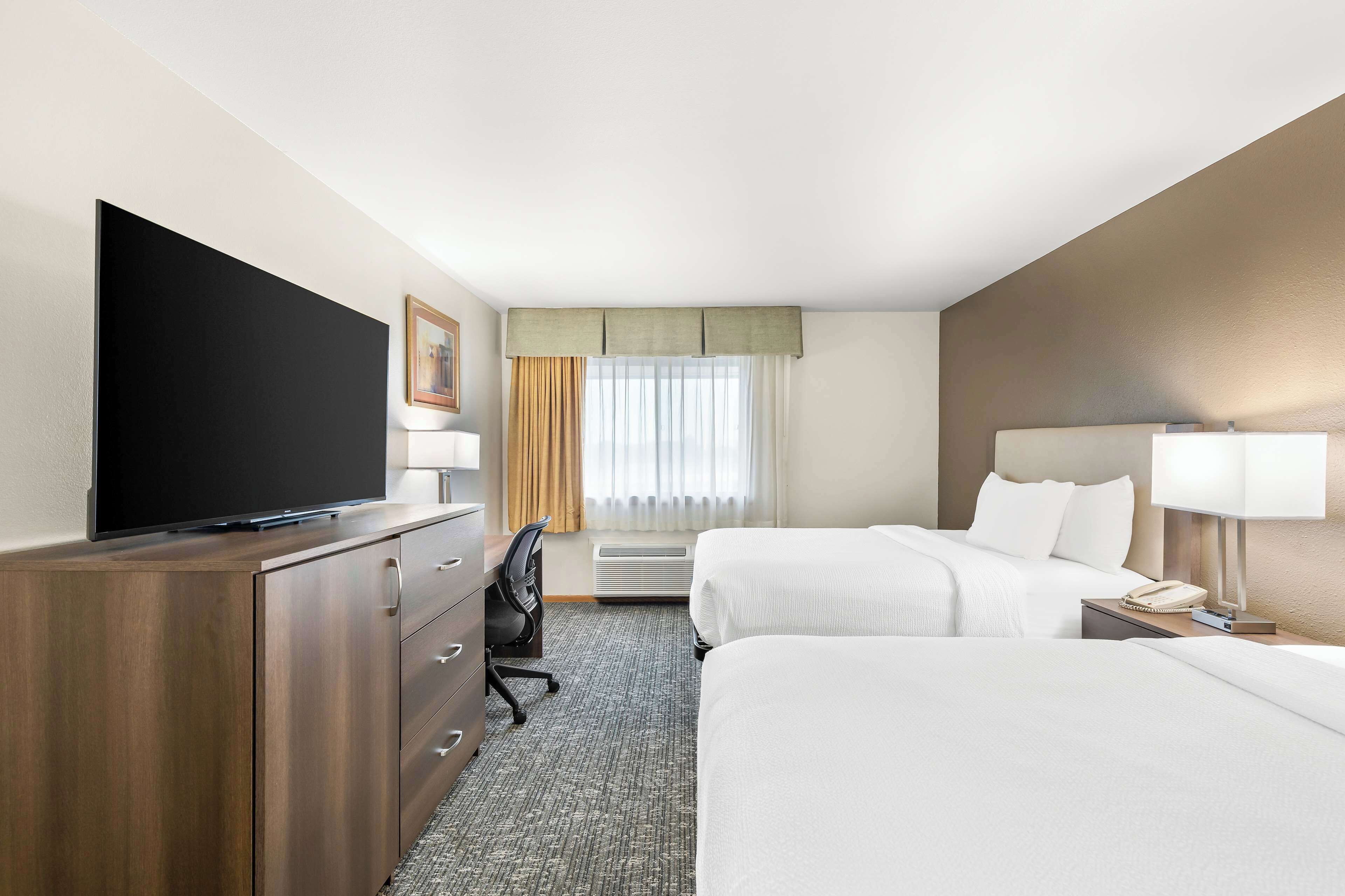 A hotel room with a flat-screen TV, two beds, a desk with a chair, a lamp, and a telephone.