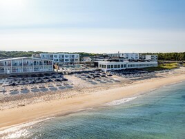 A beachfront hotel with multiple buildings, a sandy beach with sun loungers, and a clear view of the ocean.