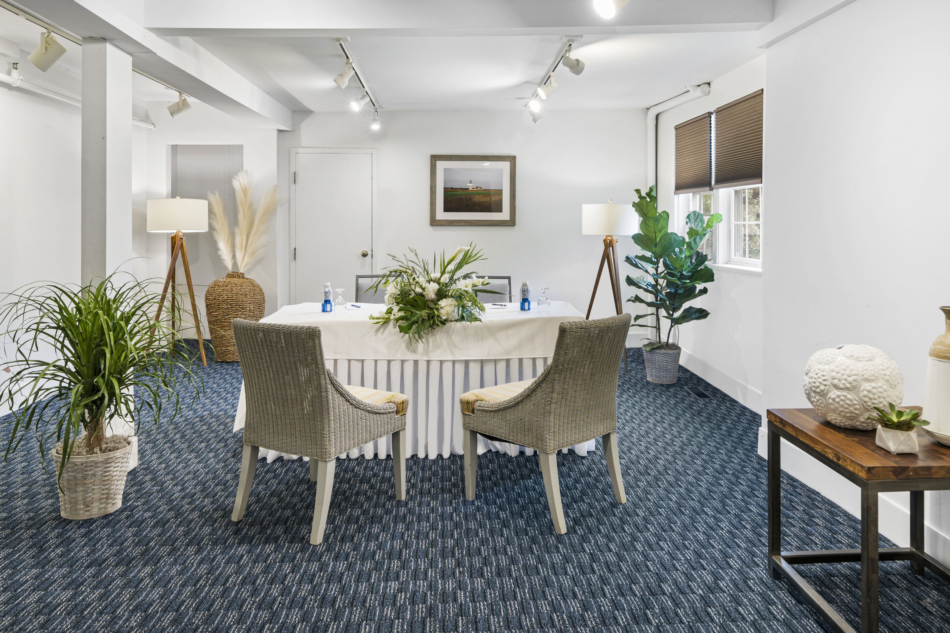 A conference room with a round table, two wicker chairs, a potted plant, a lamp, and a framed picture on the wall.