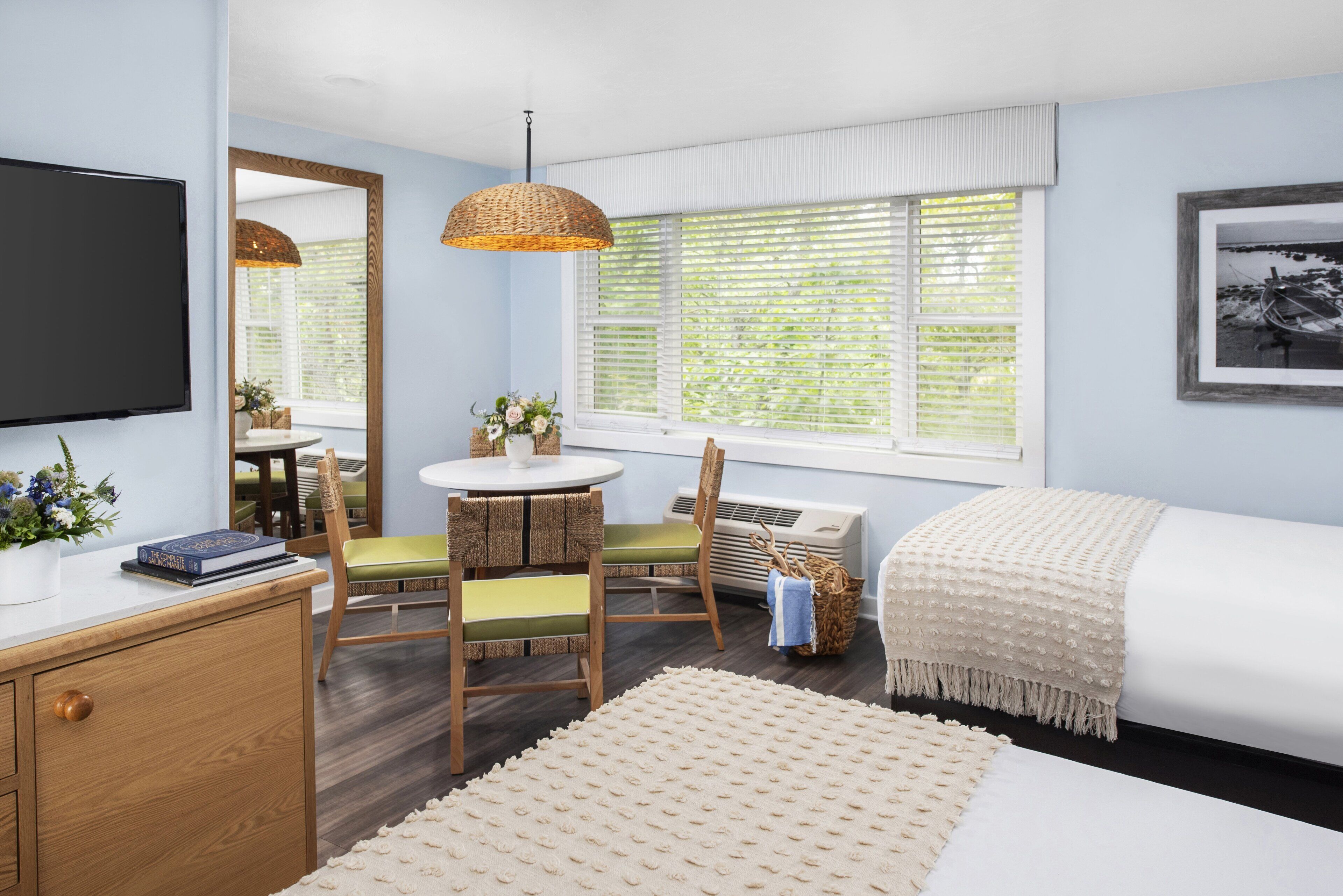 A bedroom with a bed, a desk, a chair, a TV, a window with blinds, and a mirror.