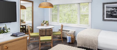 A bedroom with a bed, a desk, a chair, a TV, a window with blinds, and a mirror.