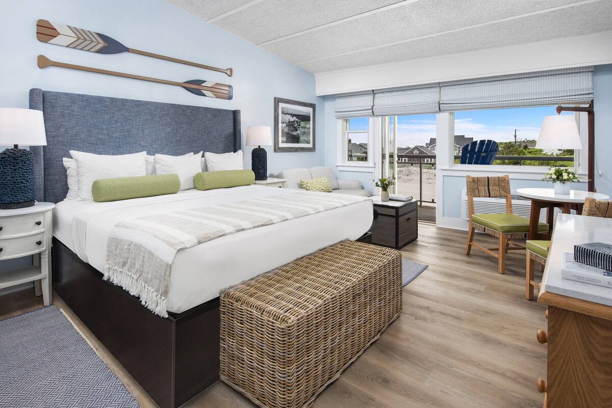 Ein Schlafzimmer mit einem großen Bett, Nachttischen, einer Korbsitzbank, einem Essbereich mit Tisch und Stühlen sowie Blick auf den Strand durch das Fenster.