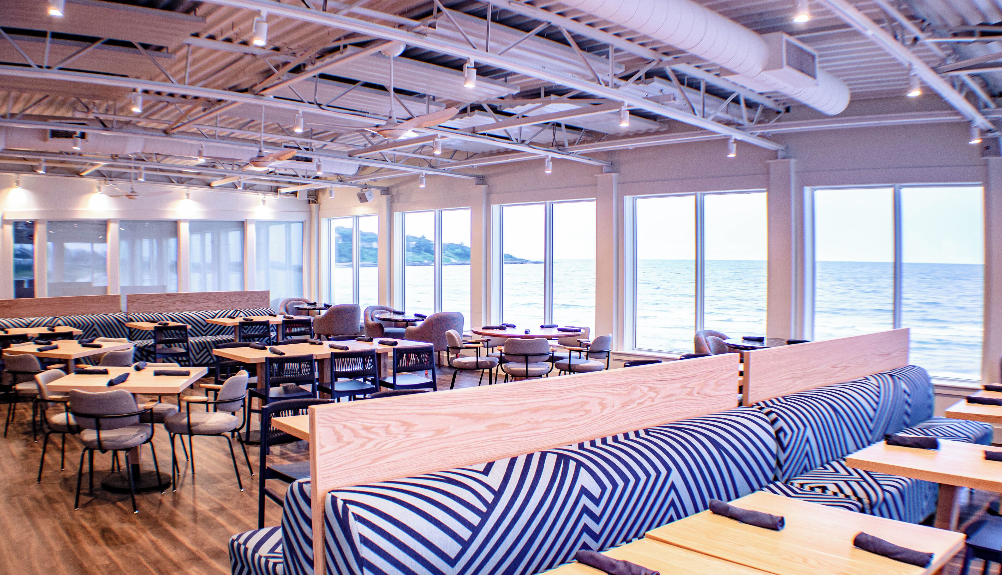 A restaurant with high ceilings, exposed ductwork, and large windows overlooking the sea.