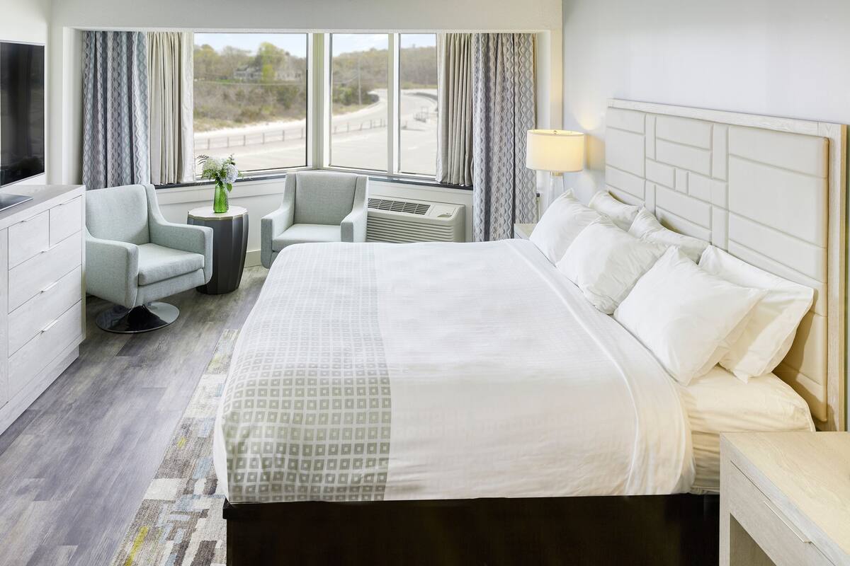 Ein Hotelzimmer mit einem großen Bett, einem Schreibtisch, einem Stuhl, einem Fernseher und einem Fenster mit Vorhängen.