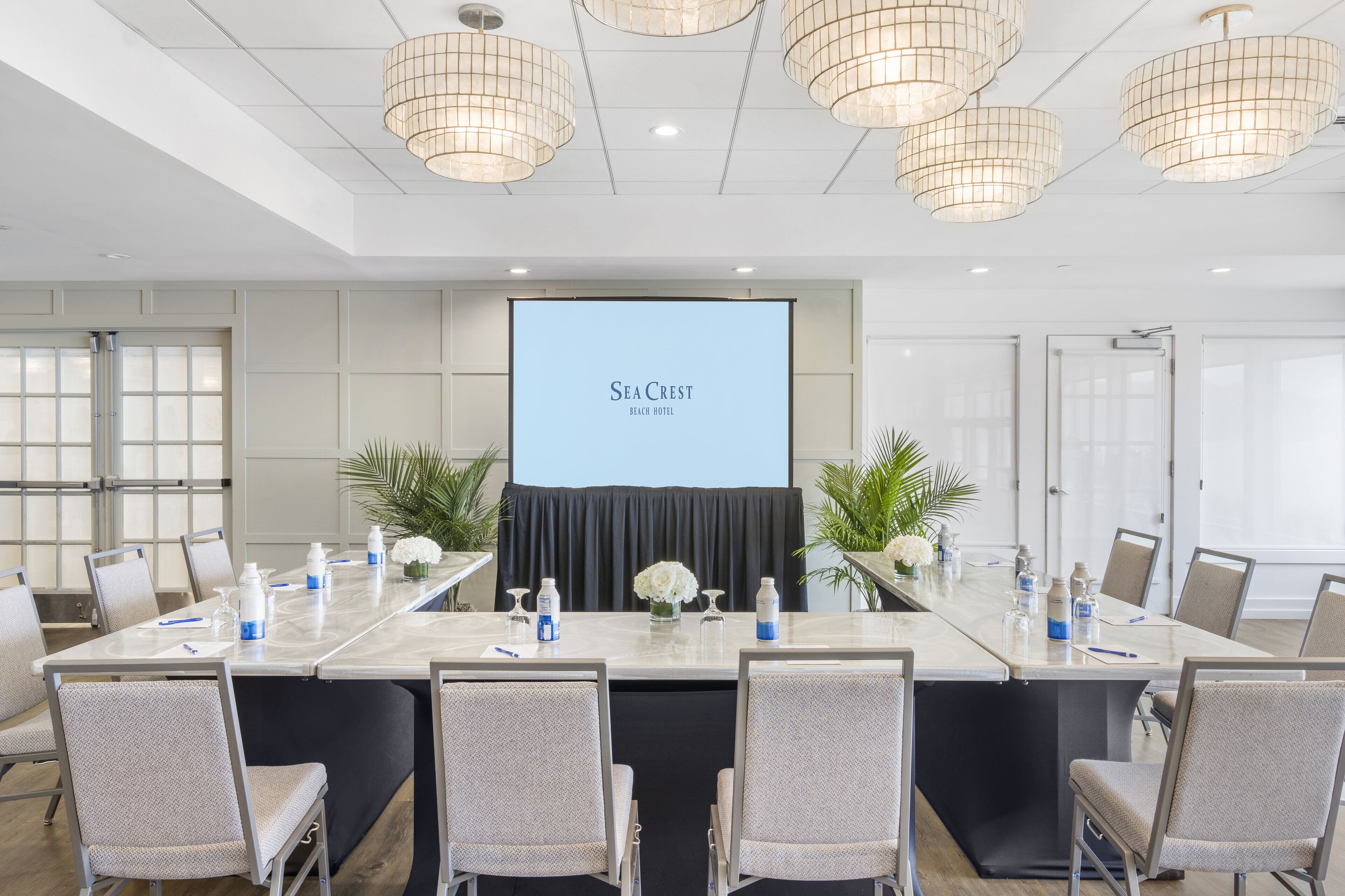A conference room with a long table, chairs, a projector screen displaying "Sea Crest Beach Hotel", and ceiling lights.