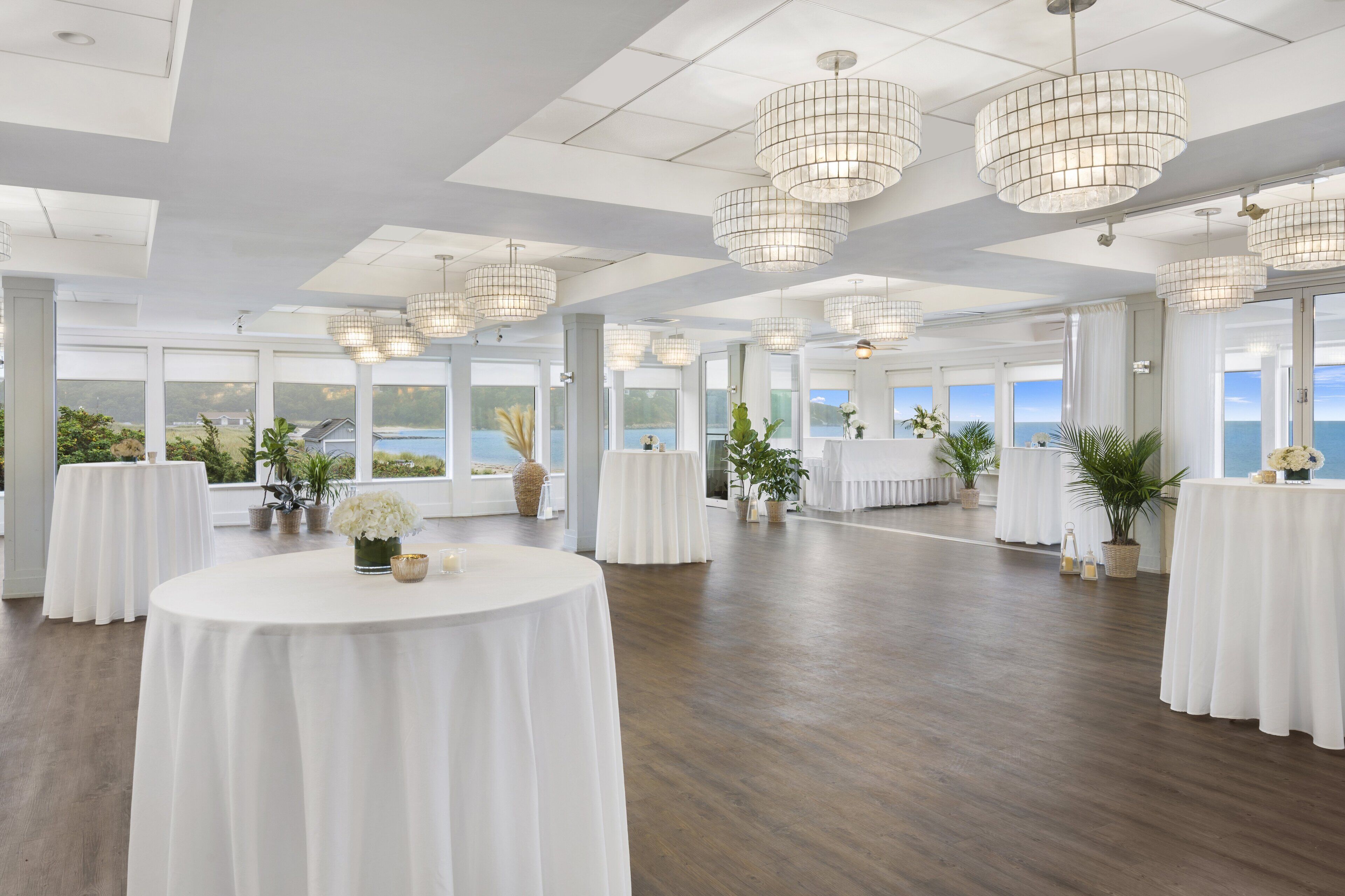 A spacious room with multiple round tables covered in white tablecloths, large windows offering a view of the sea, and modern pendant lighting.