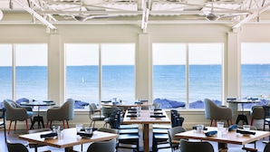 A dining area with tables and chairs, large windows with a view of the sea, and ceiling fans.