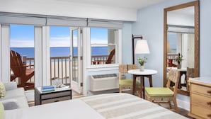 A hotel room with a bed, a desk, a chair, a lamp, a dresser, and a large mirror. There is a view of the ocean through the windows.