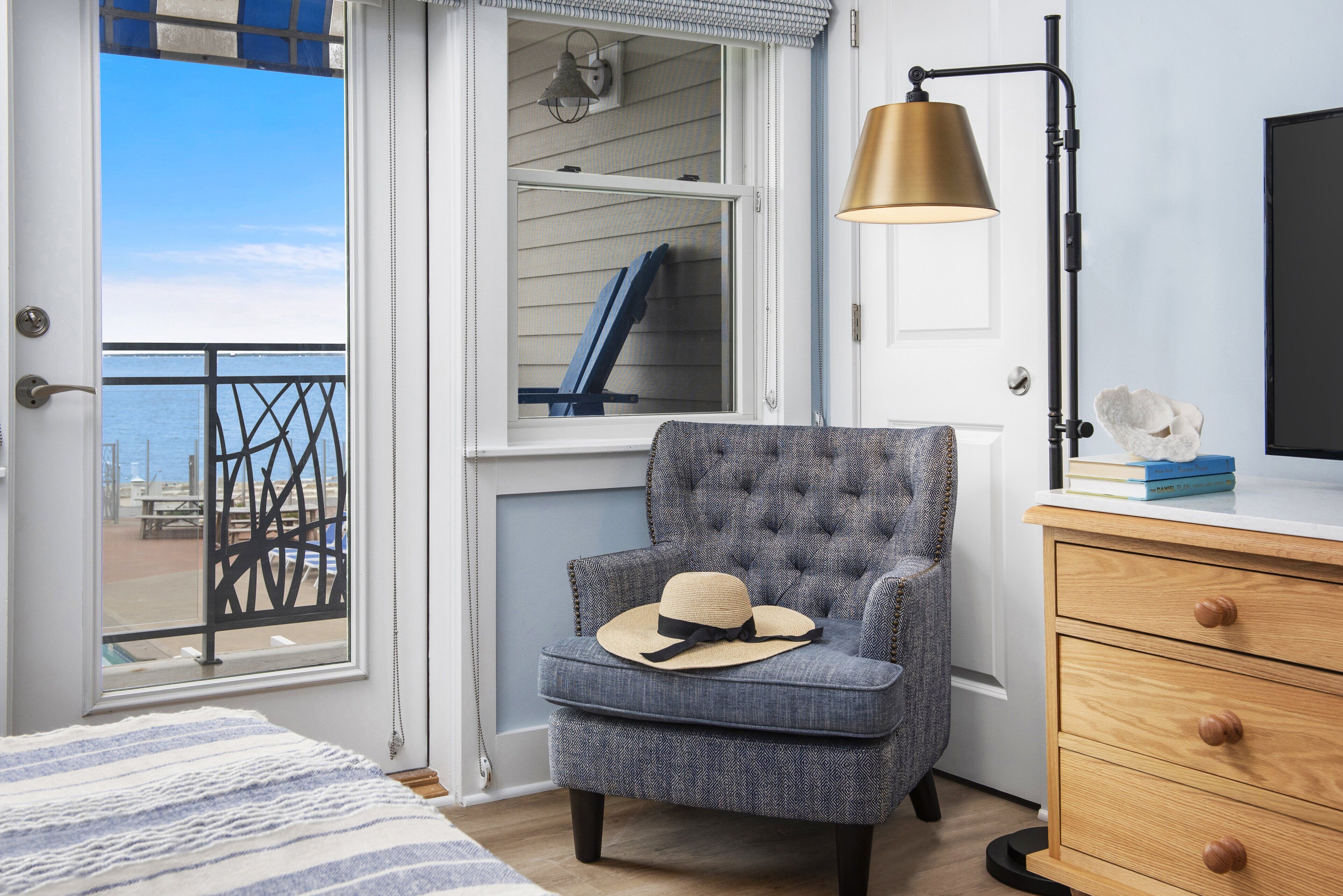 Ein Zimmer mit Meerblick, ein Sessel mit Hut, eine Lampe und eine Holzkommode.