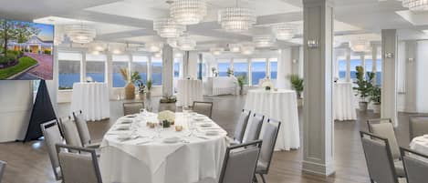 Een ruime eetruimte met ronde tafels gedekt voor een maaltijd, grote ramen met uitzicht op zee, en moderne kroonluchters.