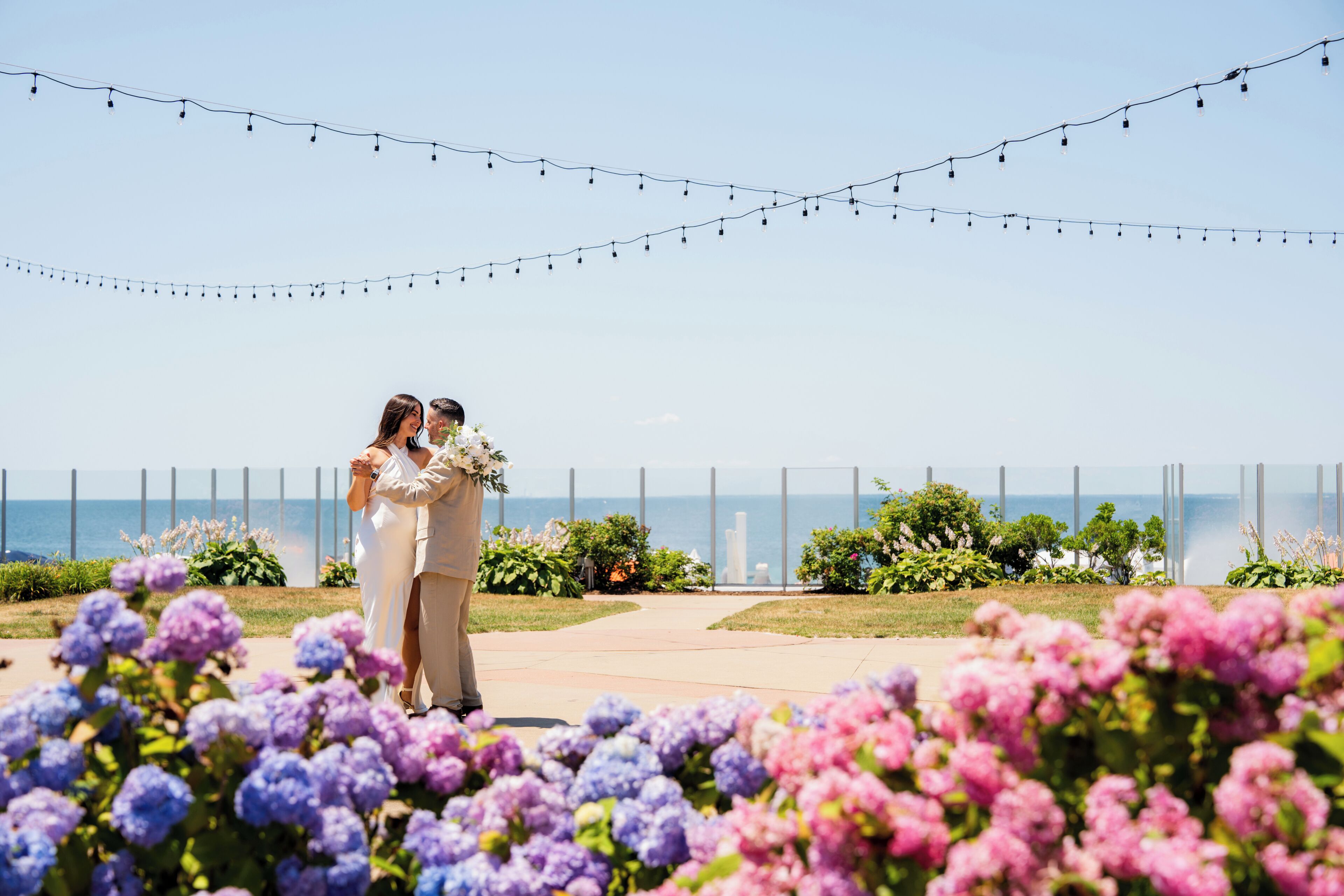 A couple standing in a garden with a view of the ocean and string lights above.