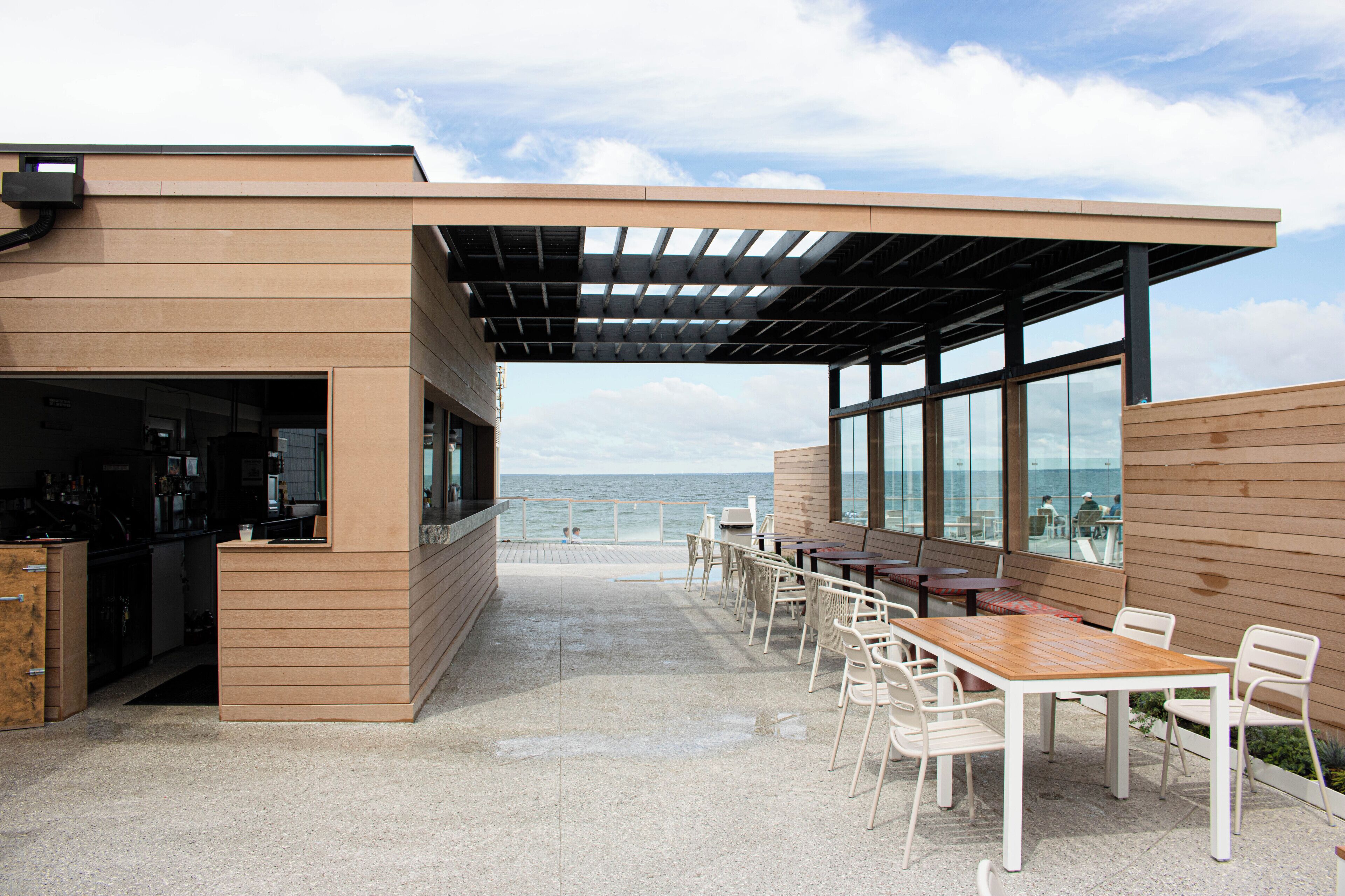 Ein moderner Essbereich im Freien mit Blick auf das Meer, mit einer Holzpergola, weißen Stühlen und einem langen Tisch.