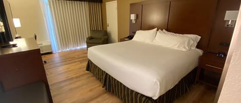 A hotel room with a large bed, wooden flooring, a desk with a computer, a chair, and a window with curtains.