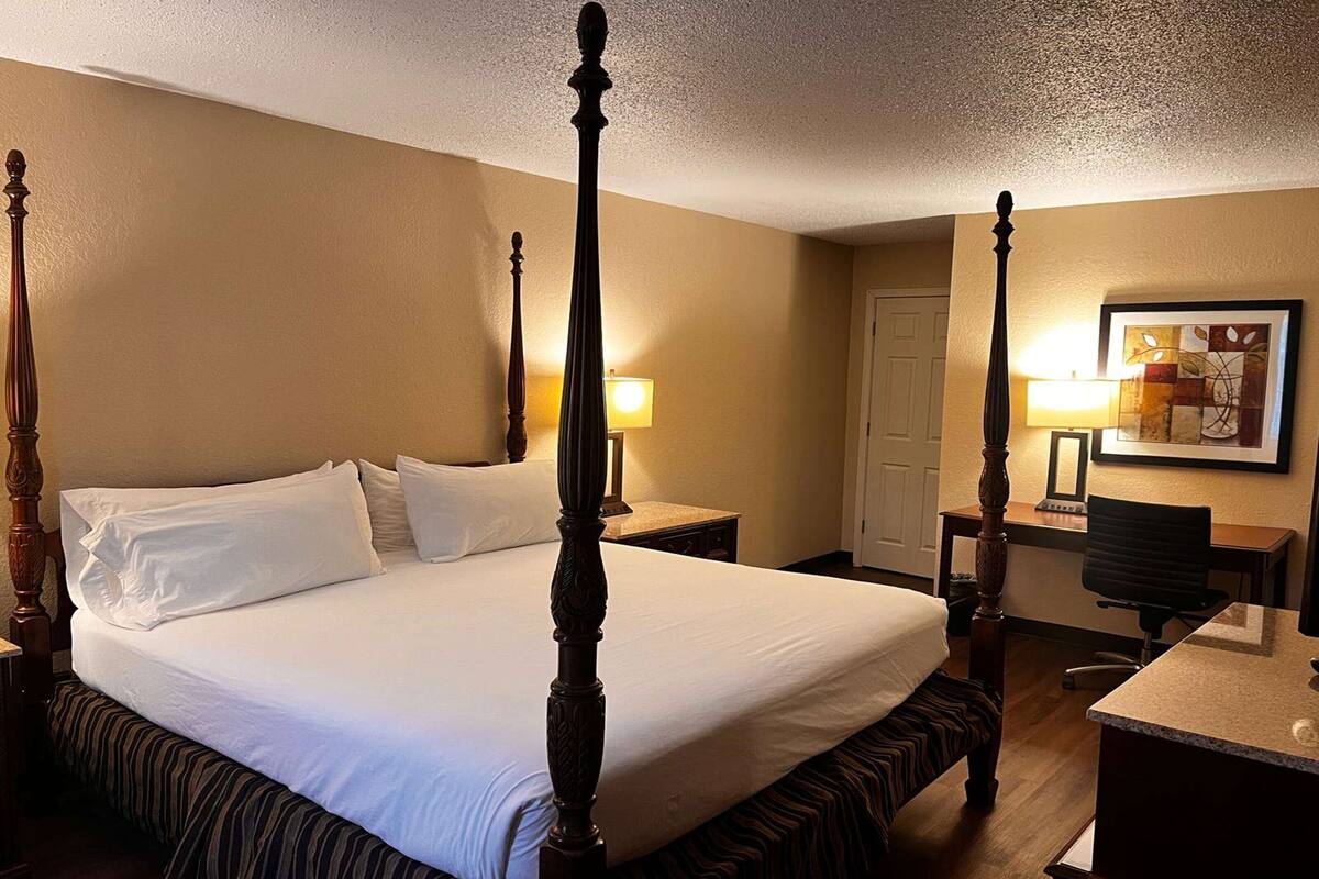 A four-poster bed with a wooden frame, a desk with a chair, and a painting on the wall.