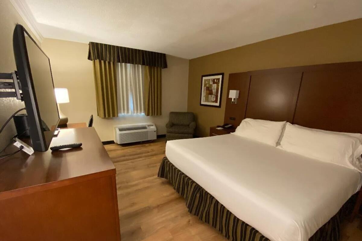 A hotel room with a large bed, a desk with a computer, a chair, a window with curtains, and a painting on the wall.