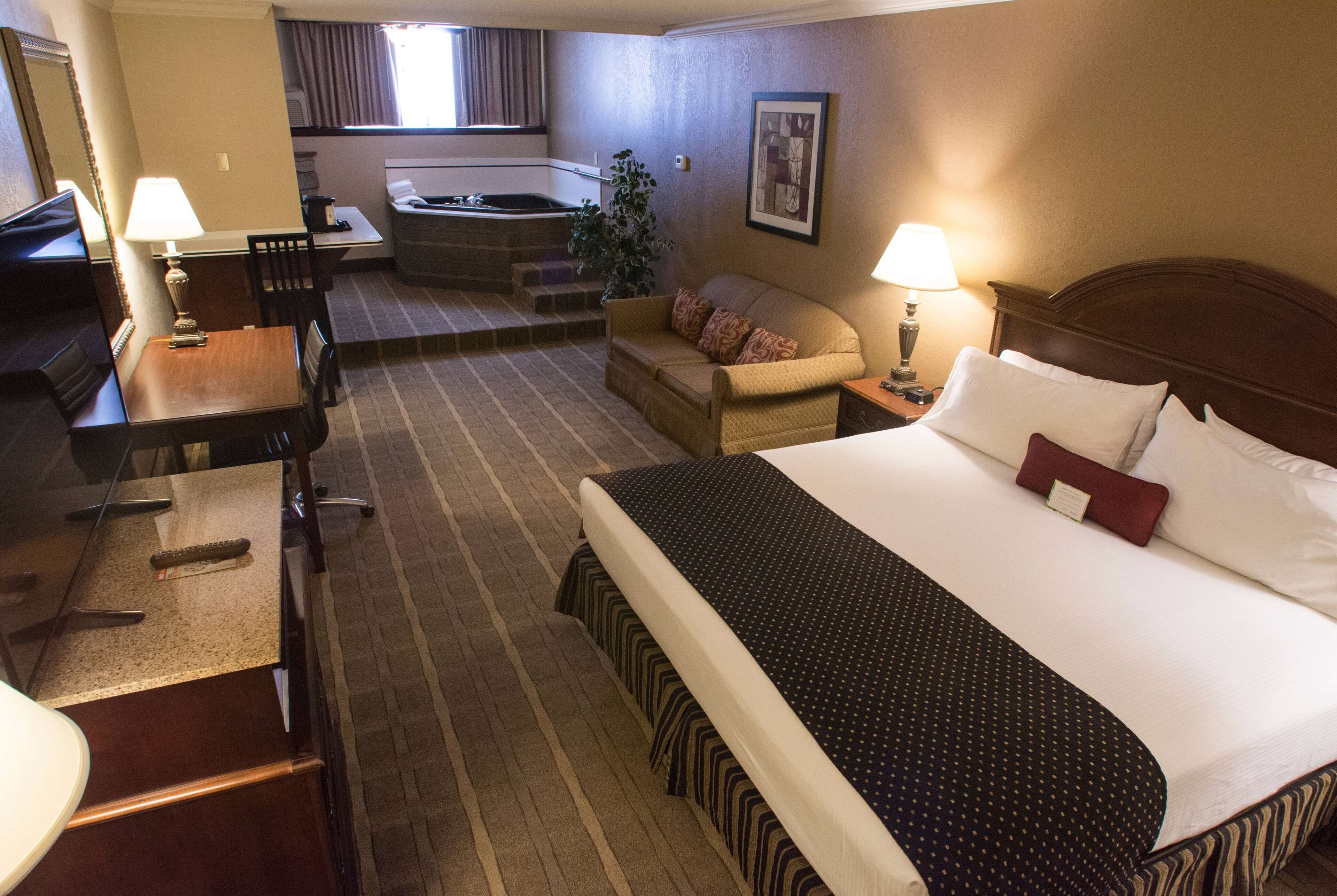 Habitación de hotel con cama, escritorio, silla, sofá, una mesita, lámpara, un cuadro y un jacuzzi al fondo.