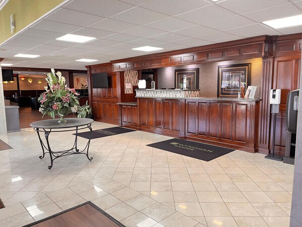 A reception area with a wooden reception desk, a vase of flowers, a tiled floor, and a black doormat with the hotel name. - Wyndham Garden Warsaw (Warsaw)