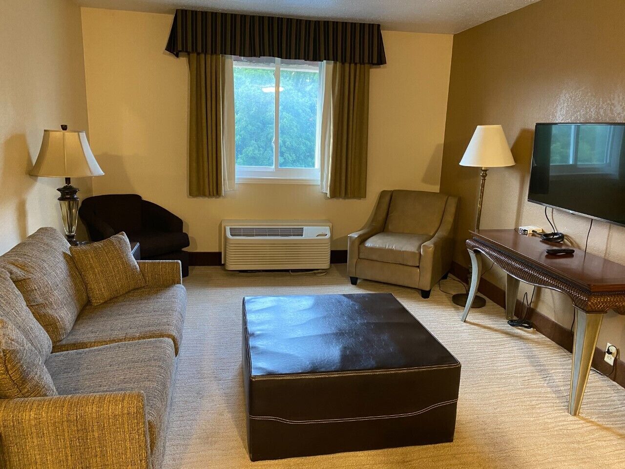 A hotel room with a sofa, two armchairs, a coffee table, a TV, and an air conditioning unit.