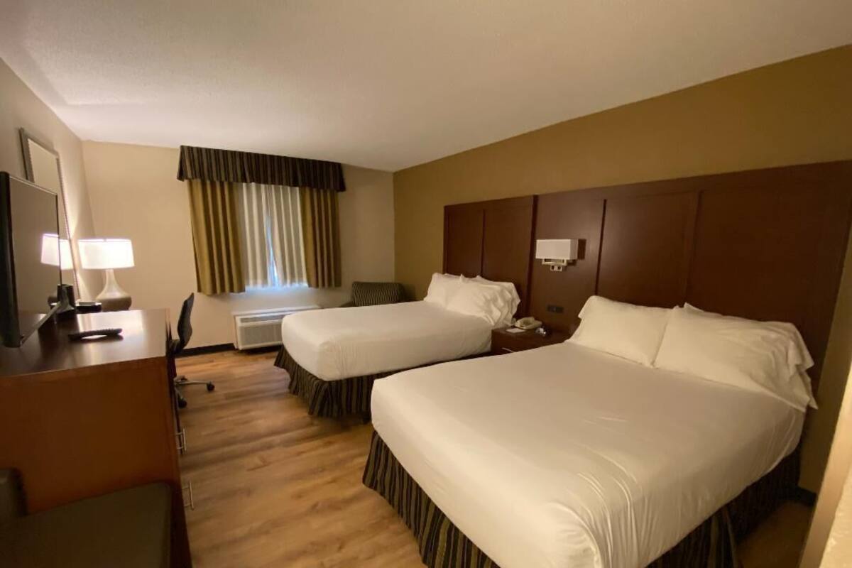 A hotel room with two beds, a desk with a lamp, a chair, and a window with curtains.