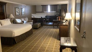 A hotel room with a large bed, a sofa, a small table, a desk, a chair, a kitchenette, and a bathroom.