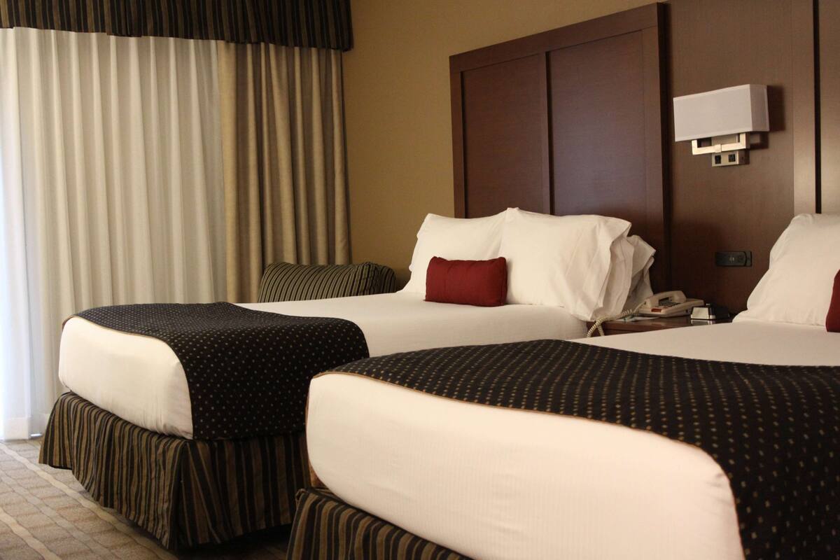 A hotel room with two beds, a large window with curtains, a bedside table with a phone, and a wall-mounted light.