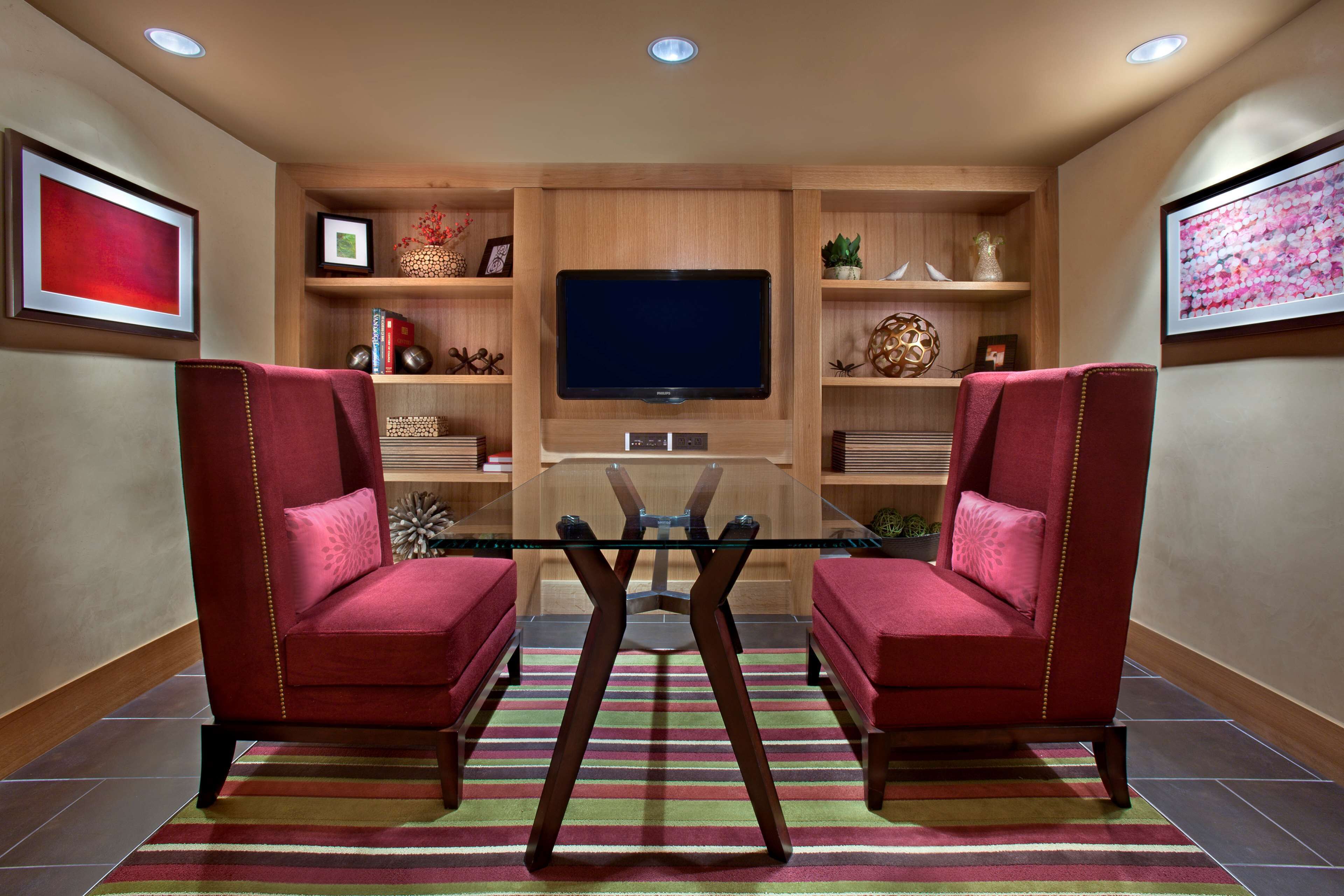 Een moderne woonkamer met twee rode fauteuils, een glazen salontafel, een flatscreen televisie, houten planken met decoratieve objecten en een gestreept vloerkleed.