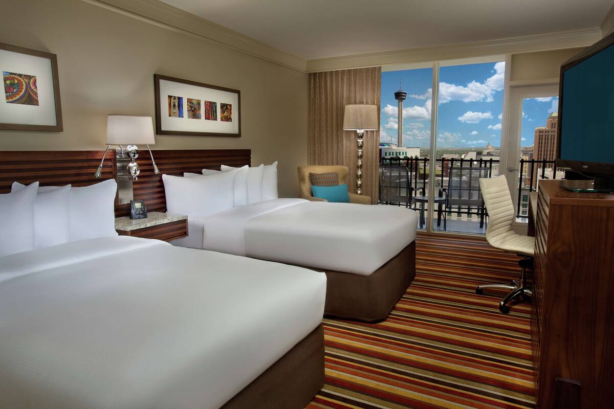 A hotel room with two beds, a desk, a chair, a TV, and a balcony with a city view.