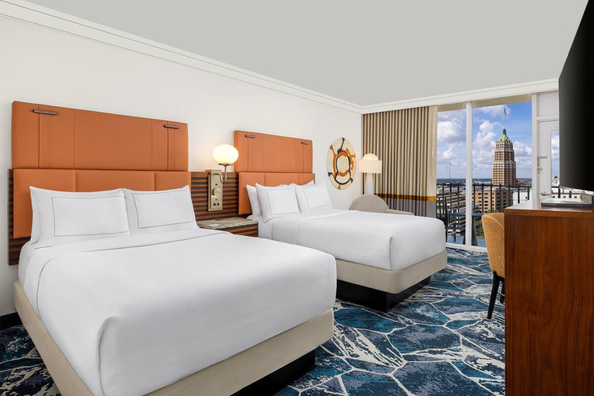 A hotel room with two beds, a desk, a TV, and a large window with a city view.