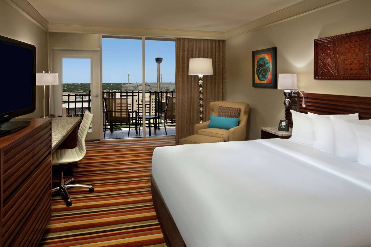 A hotel room with a bed, a desk, a chair, a television, and a view of a tower through the balcony doors.