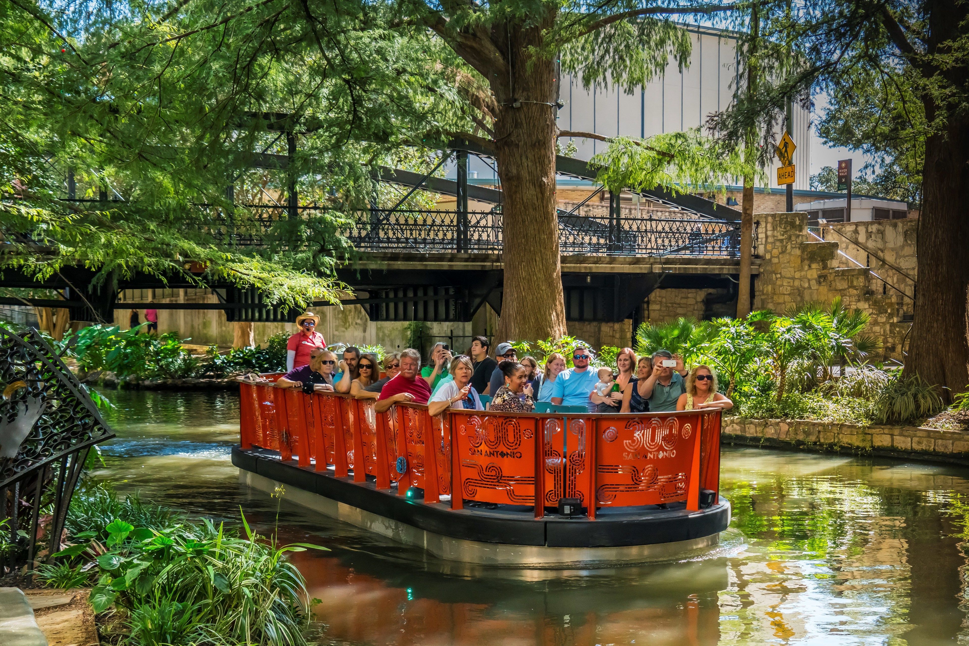 A boat ride with passengers, surrounded by lush greenery and a stone bridge in the background.