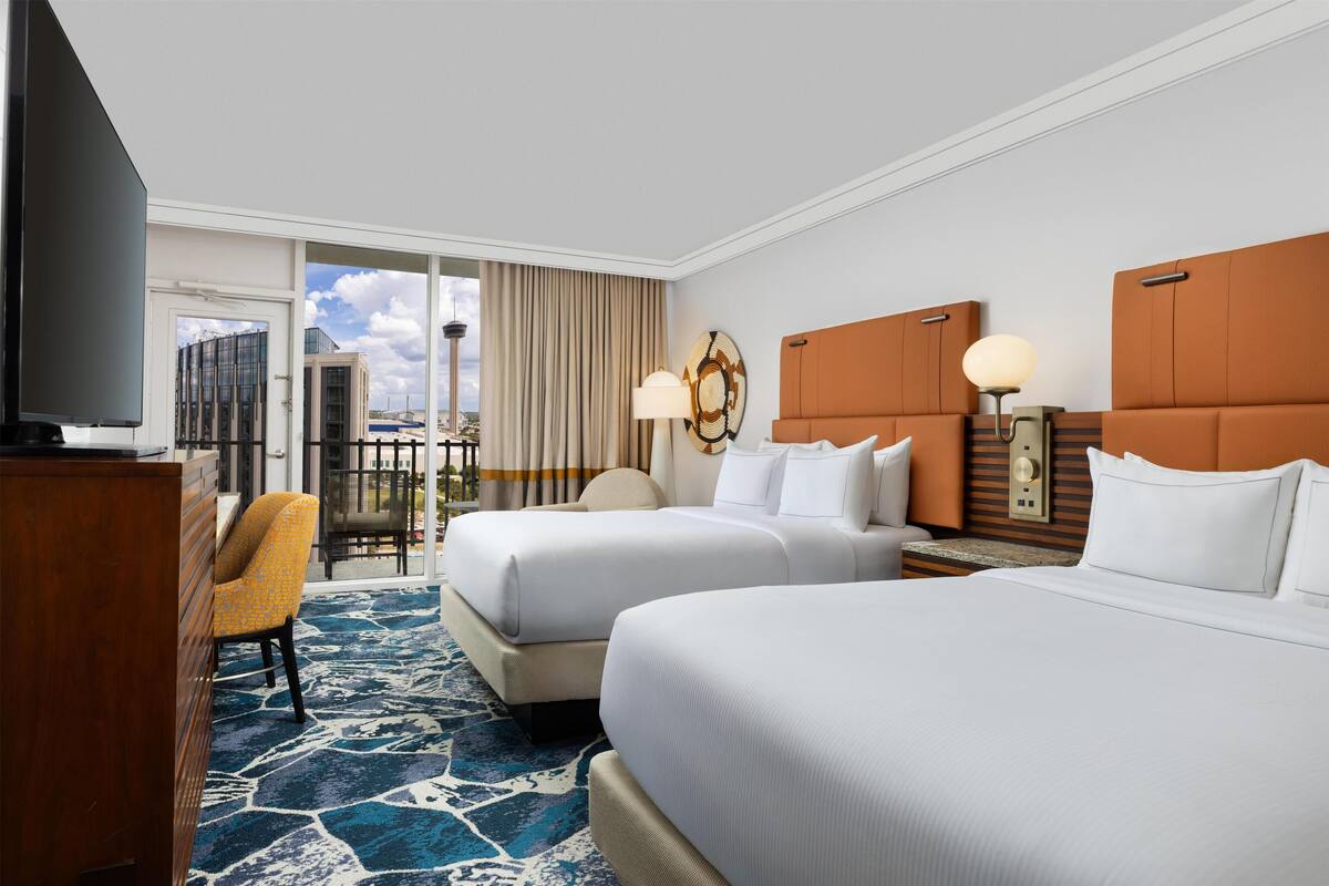 A hotel room with two beds, a television, a desk, and a view of the cityscape.