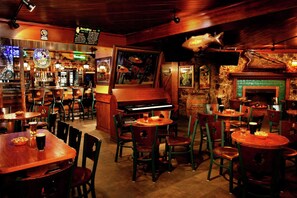 A cozy bar area with wooden tables and chairs, a piano, and a stone wall with a fireplace.