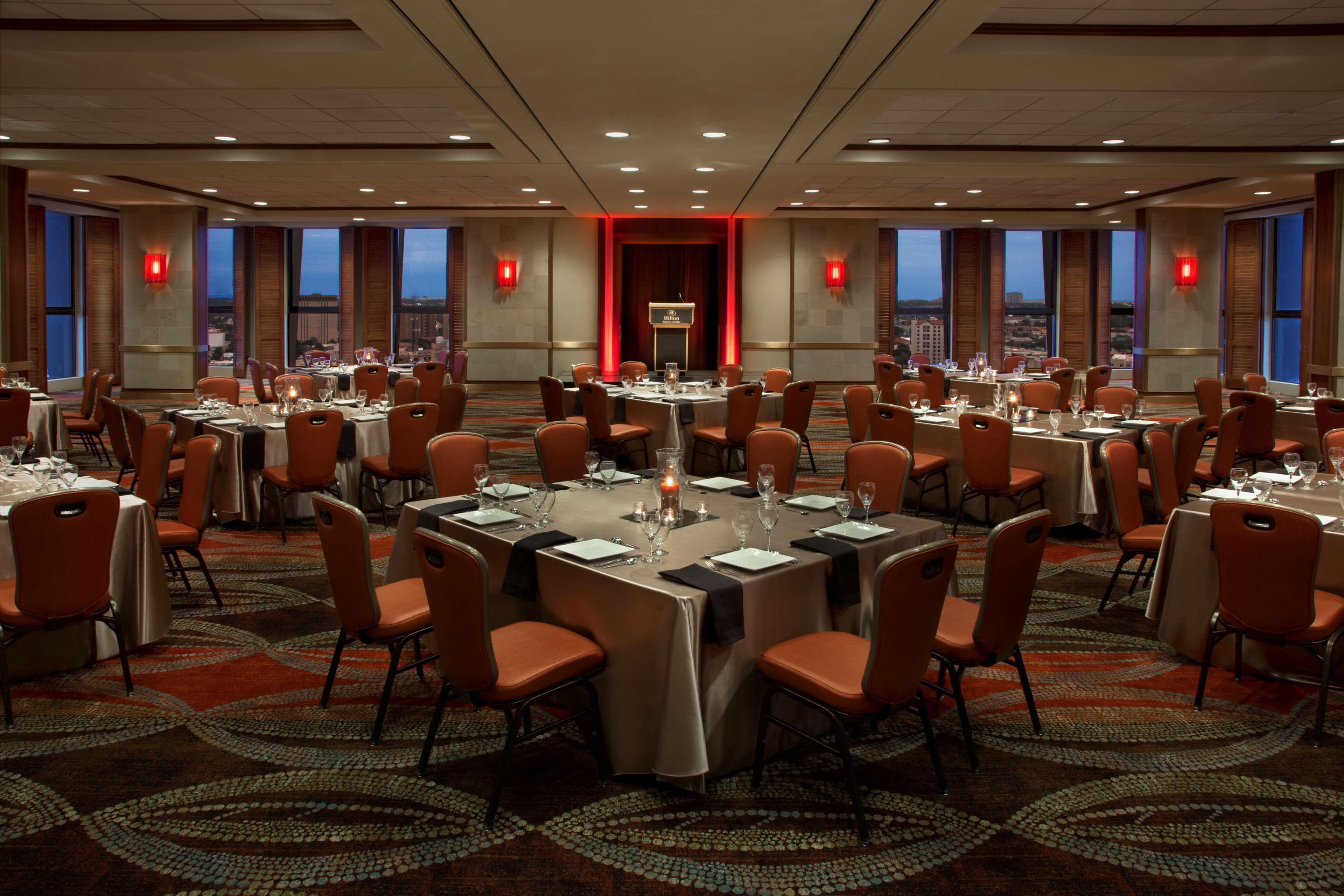 A large conference room with multiple round tables, each set with chairs and tableware, featuring a city view through large windows.