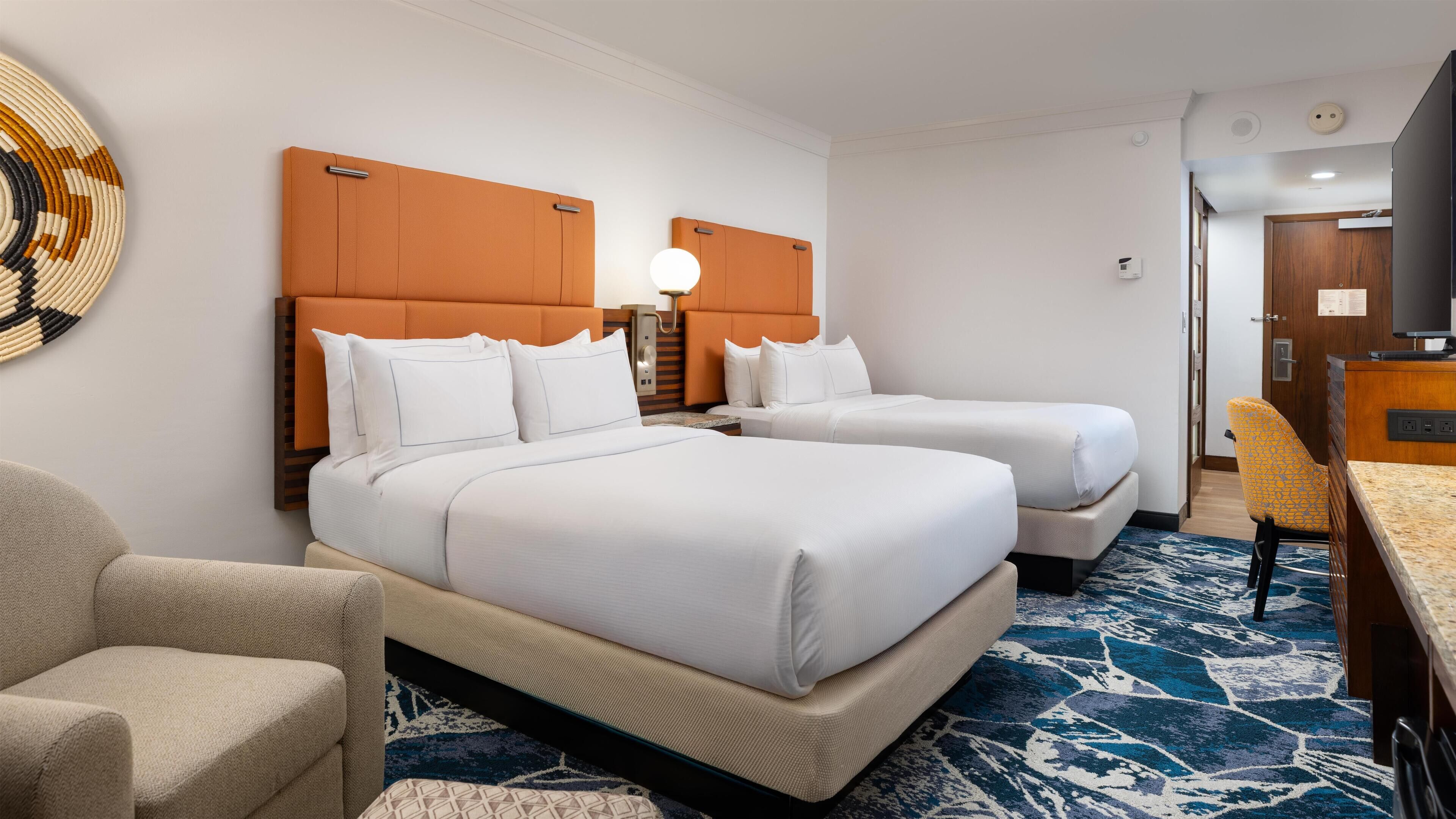 A hotel room with two beds, a desk, a chair, and a patterned carpet.