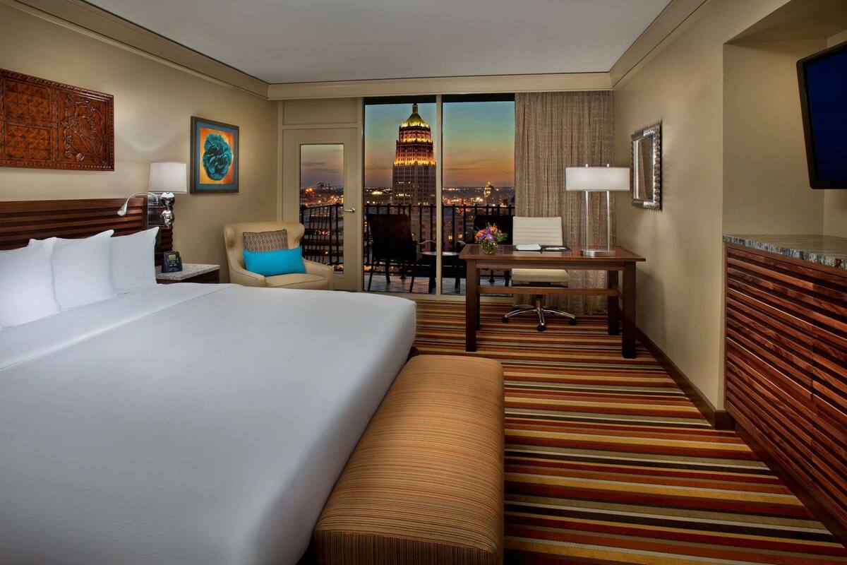A hotel room with a large bed, a desk, a chair, a bench, and a view of a cityscape.