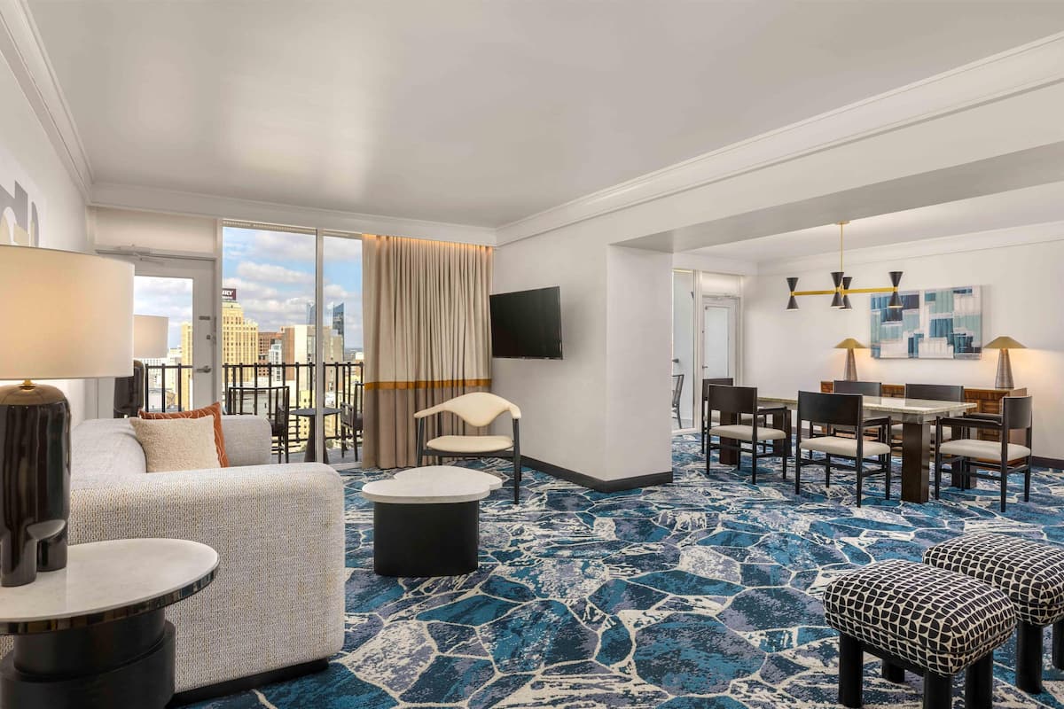 A hotel room with a large window, a sofa, a dining table with chairs, a TV mounted on the wall, and a patterned carpet.