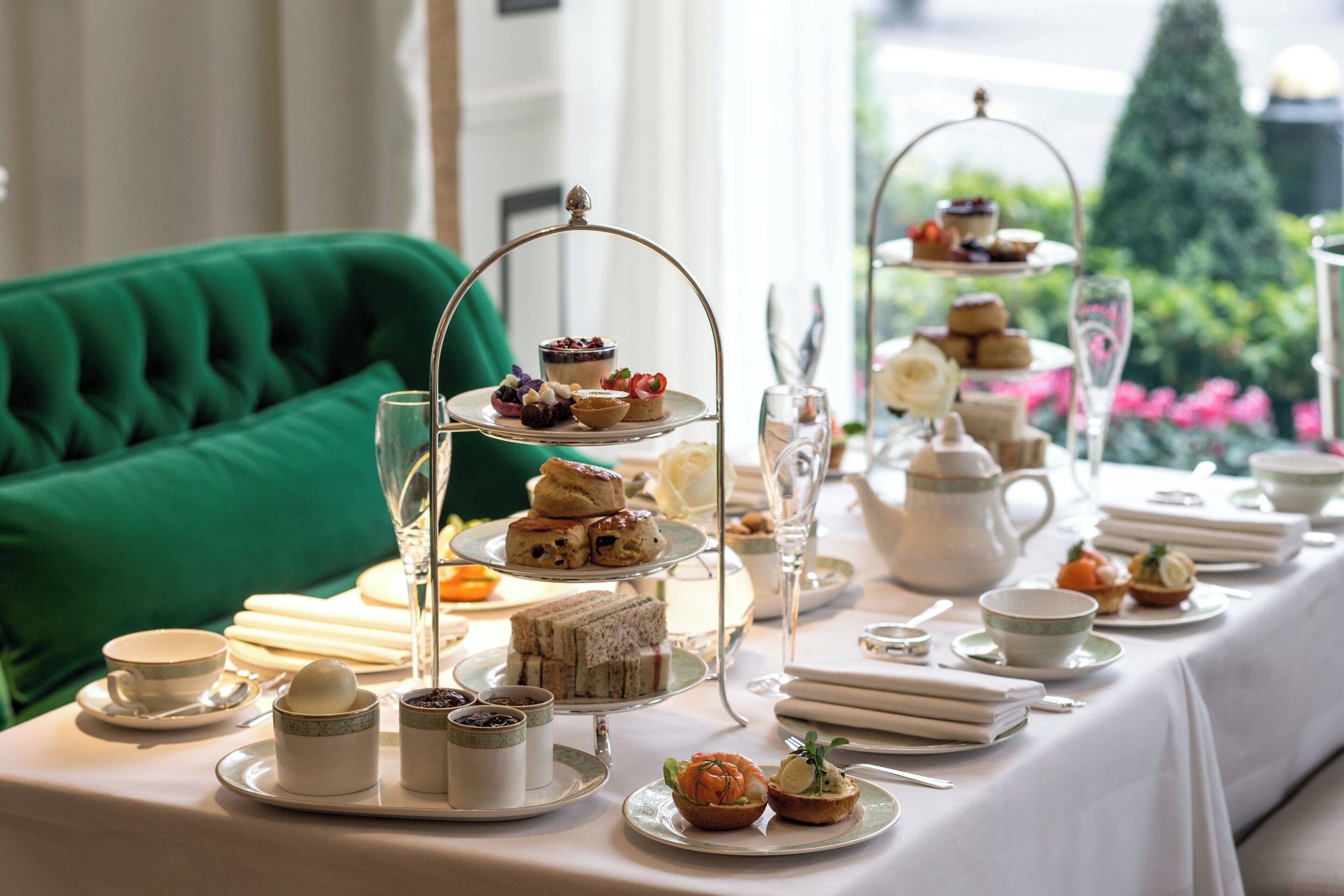 A table set for a tea service with multiple tiers of pastries and sandwiches, tea cups, and a green upholstered chair in the background.