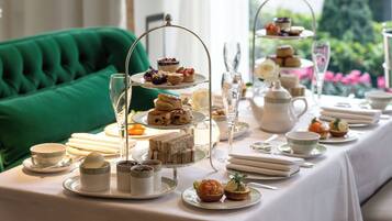 A table set for a tea service with multiple tiers of pastries and sandwiches, tea cups, and a green upholstered chair in the background.