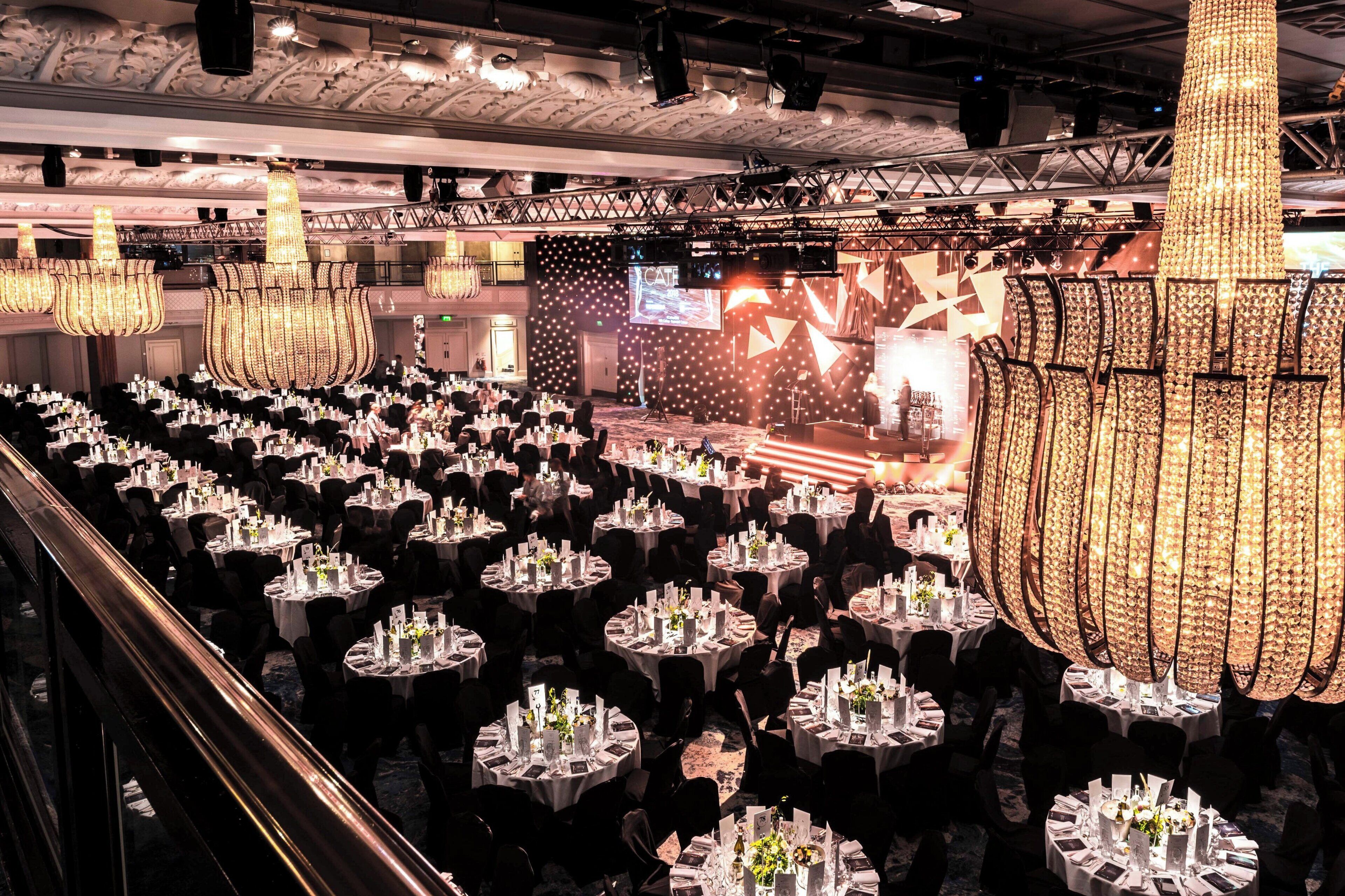 A banquet hall with round tables set for a formal event, featuring a stage with a large screen and decorative lighting.