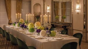Une longue table à manger, dressée pour un repas formel, agrémentée de nombreuses bougies, de compositions florales et de chaises recouvertes d’un tissu vert.