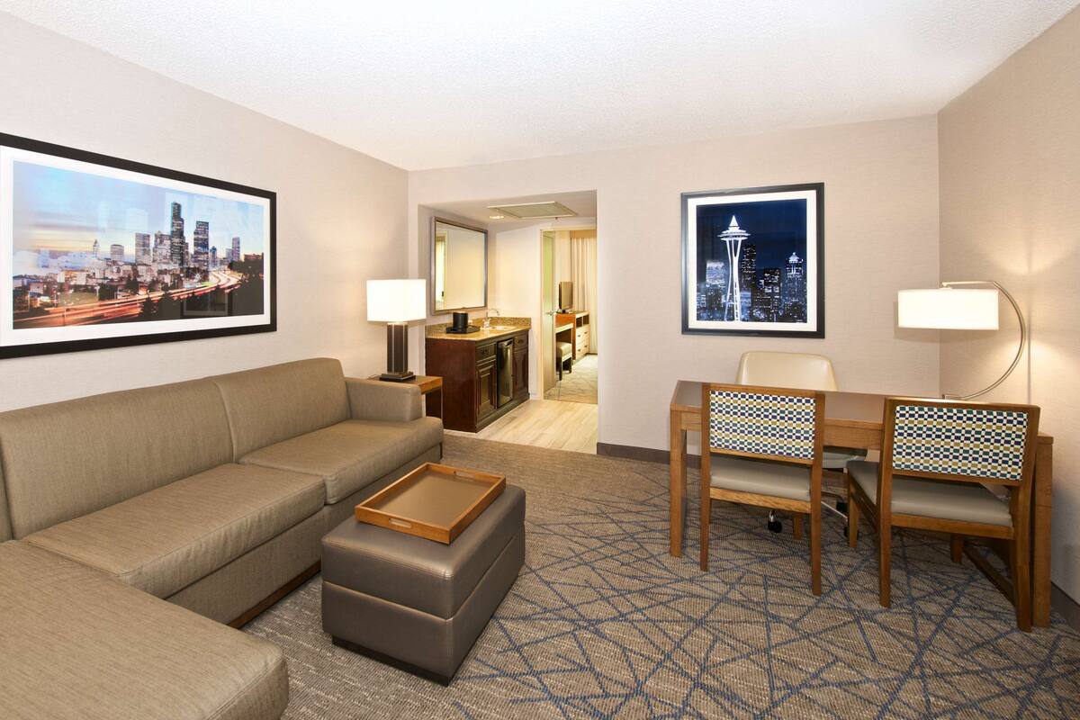 Een hotelkamer met een bank, voetenbankje, een bureau met stoel, een spiegel en twee ingelijste foto's aan de muur.