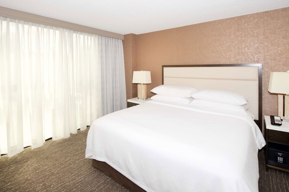 Una habitación de hotel con una cama grande, dos mesitas de noche, una lámpara en cada una y un ventanal con cortinas traslúcidas.