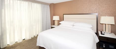 A hotel room with a large bed, two bedside tables with lamps, a nightstand with a phone, and a window with sheer curtains.