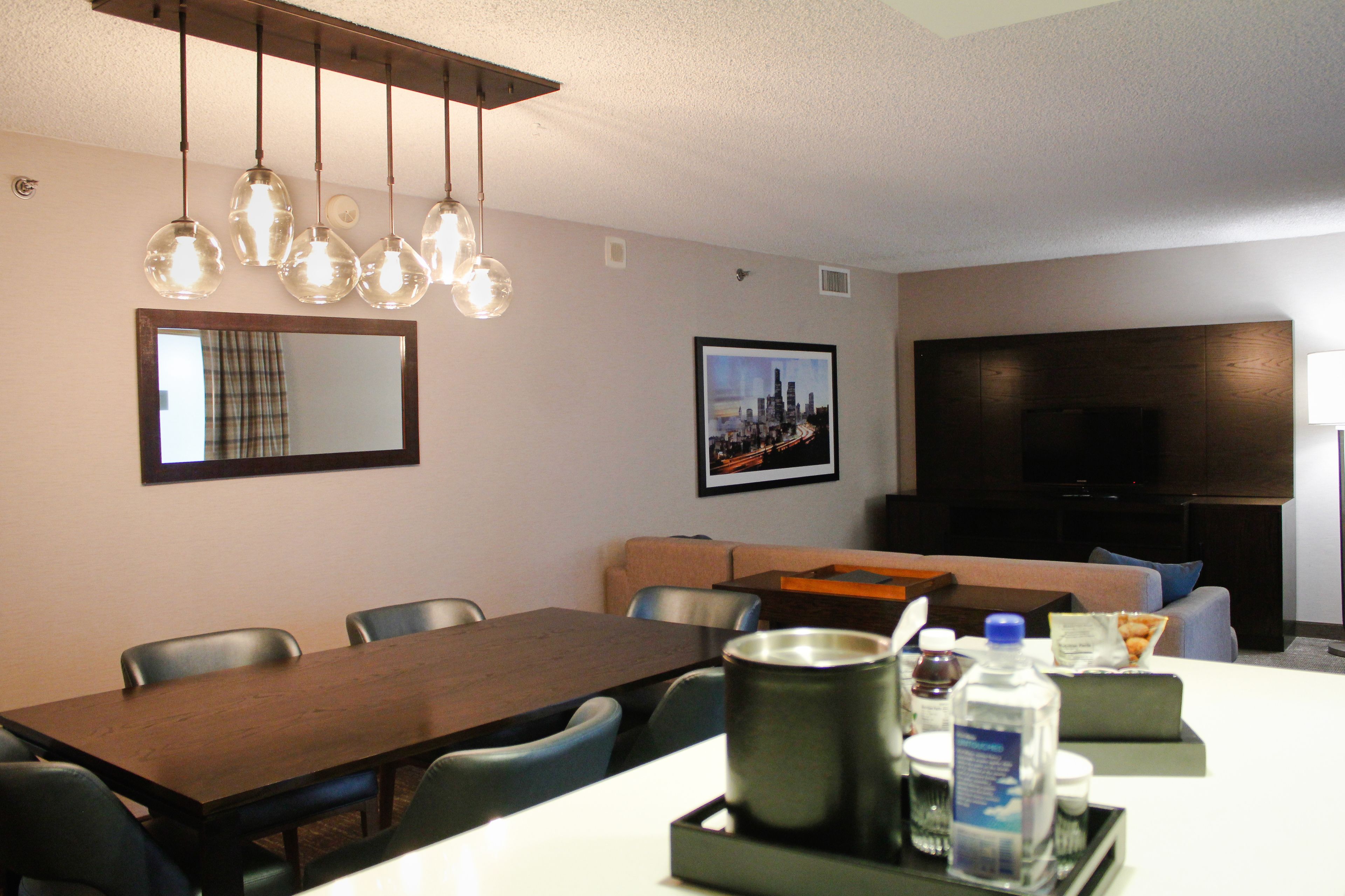 A hotel room with a dining table, chairs, a sofa, a TV, and a framed cityscape picture on the wall.