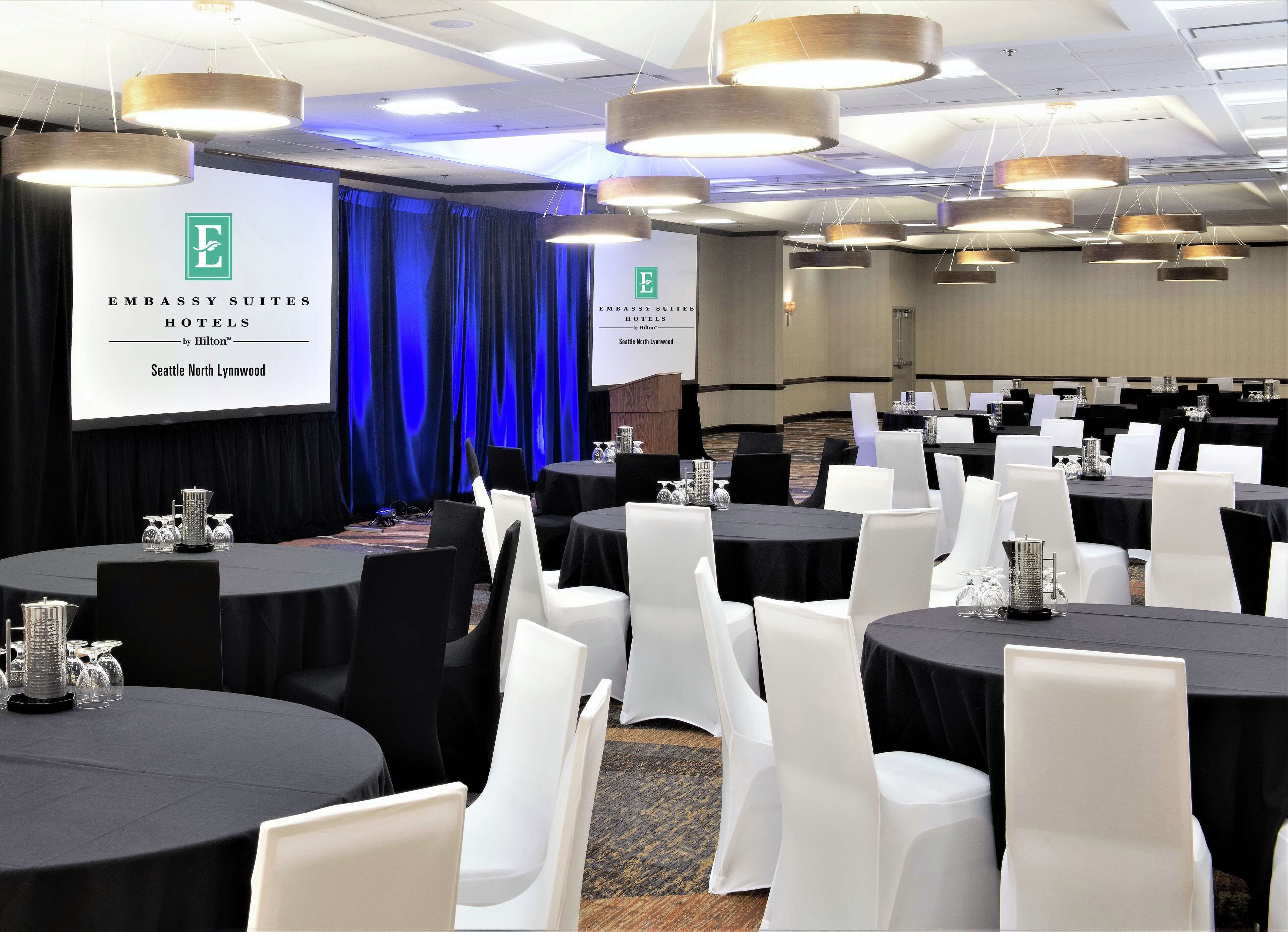 A conference room with round tables, white chairs, and a large screen displaying the logo of Embassy Suites Hotels.