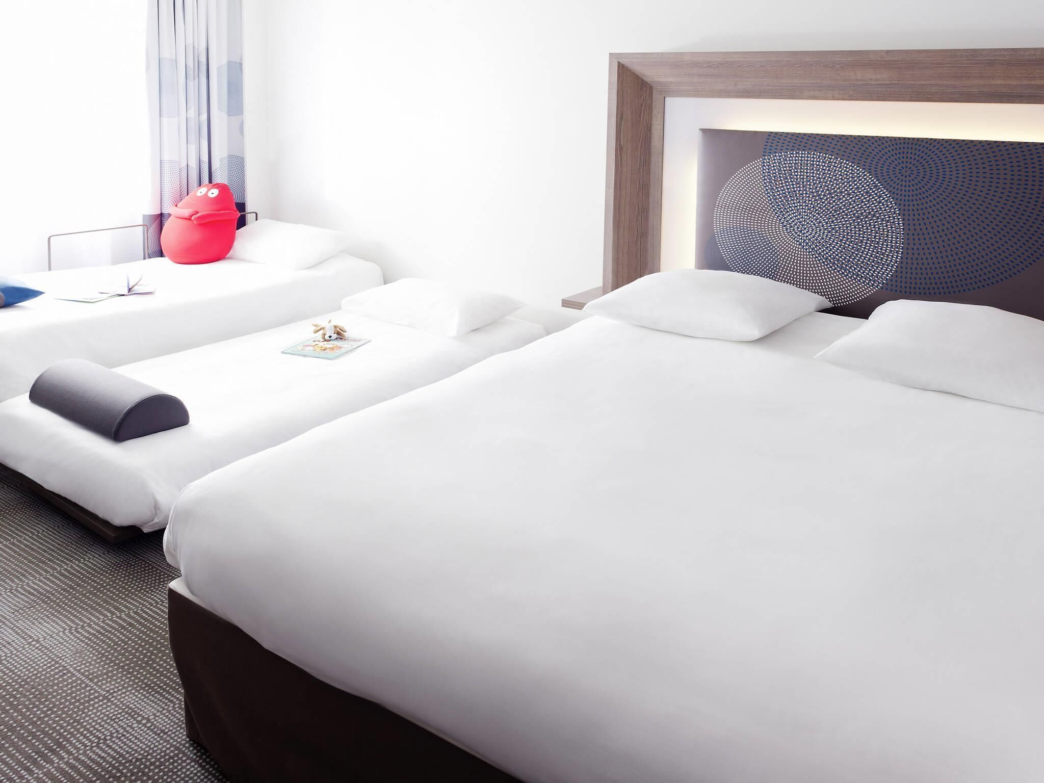 Una camera d'albergo con due letti, un giocattolo rosso sul letto a sinistra, un quadro astratto incorniciato sopra il letto a destra e una finestra con tende.