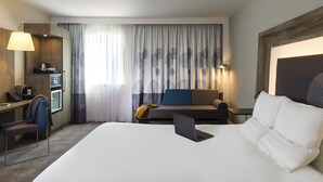 A hotel room with a bed, a desk, a chair, a sofa, a lamp, and a large window with curtains.