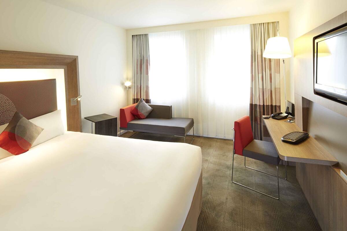 A hotel room with a bed, a desk with a telephone, a chair, a sofa, and a television.