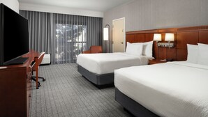 A hotel room with two beds, a desk with a TV, a chair, a lamp, and a window with curtains.