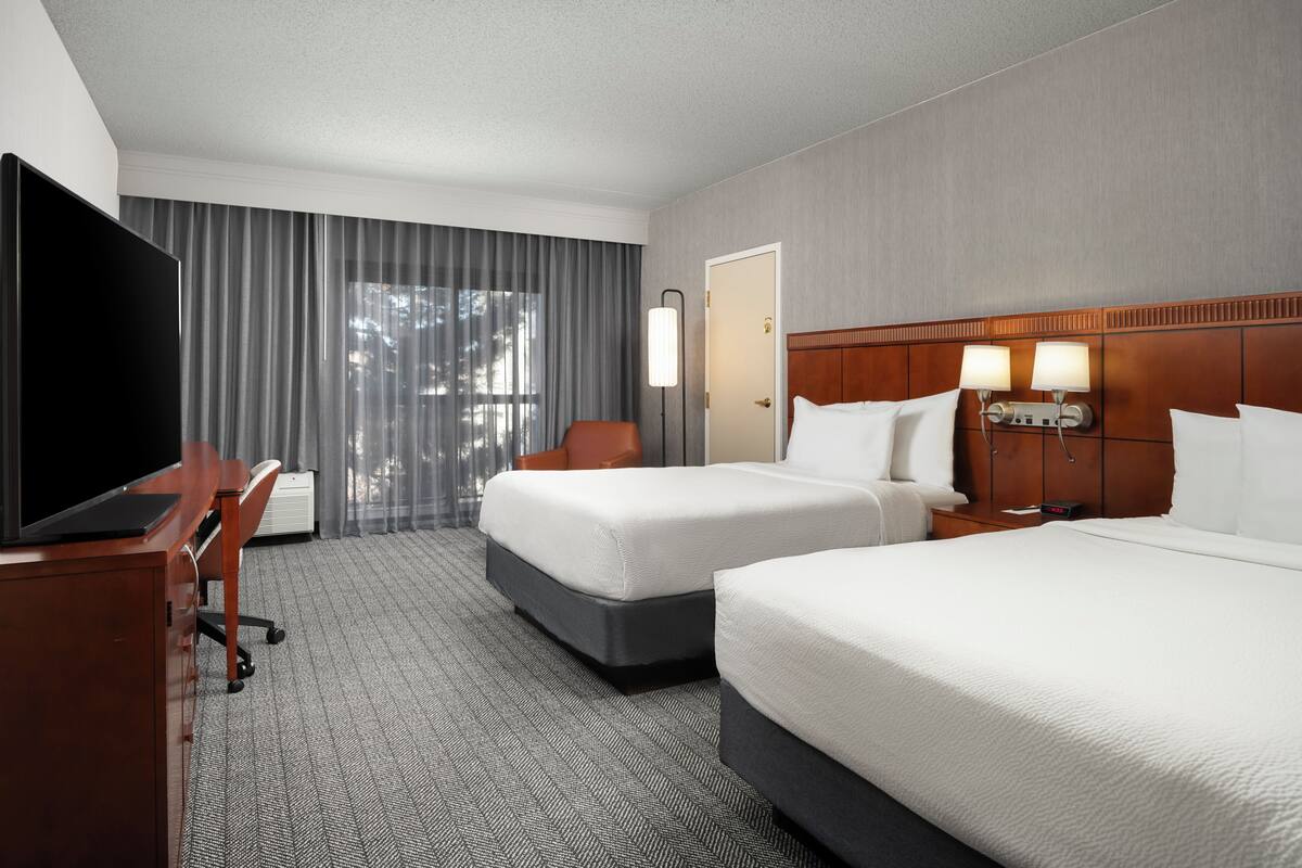 A hotel room with two beds, a desk with a TV, a chair, a lamp, and a window with curtains.